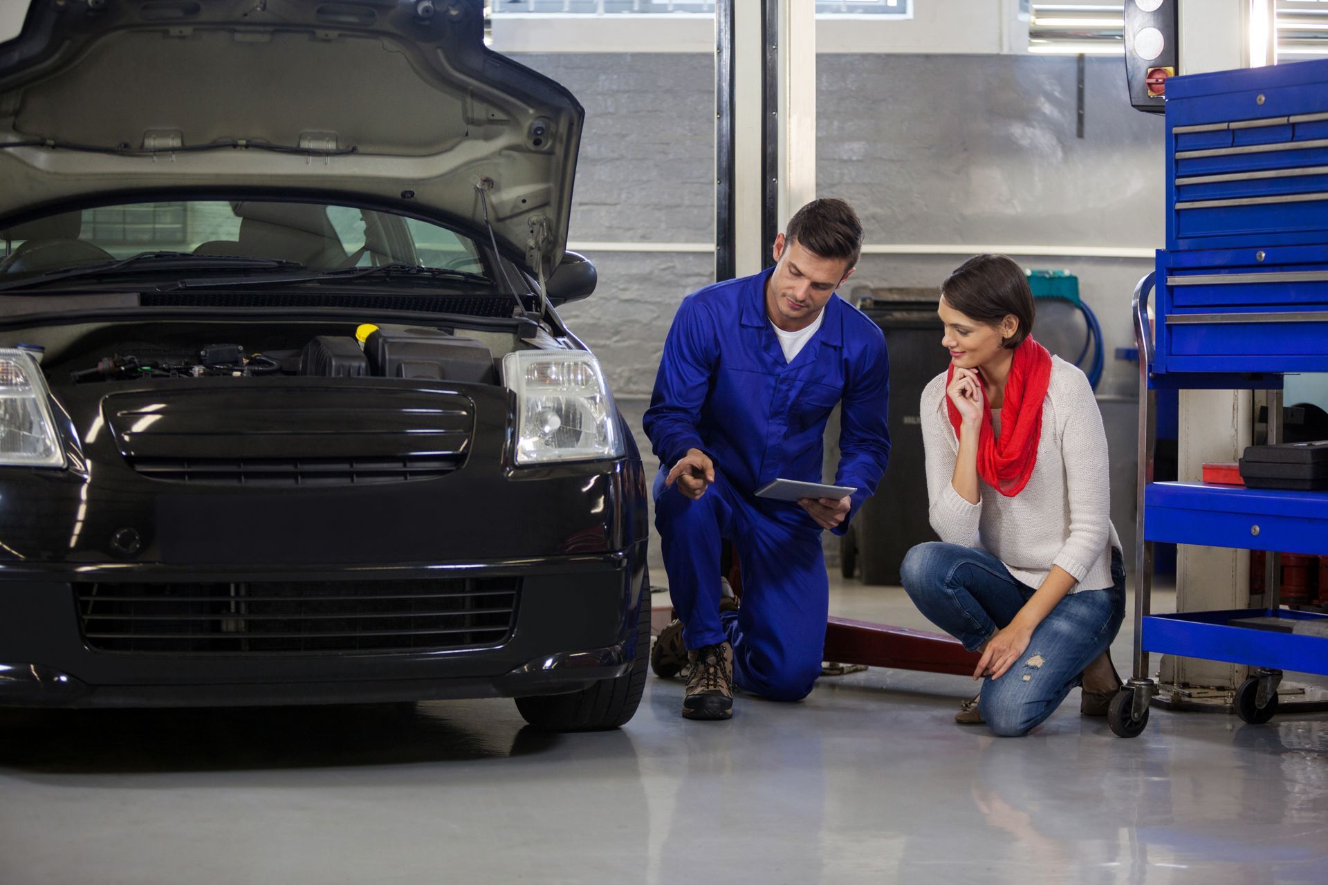 A man and a woman are looking under the hood of a car in a garage.