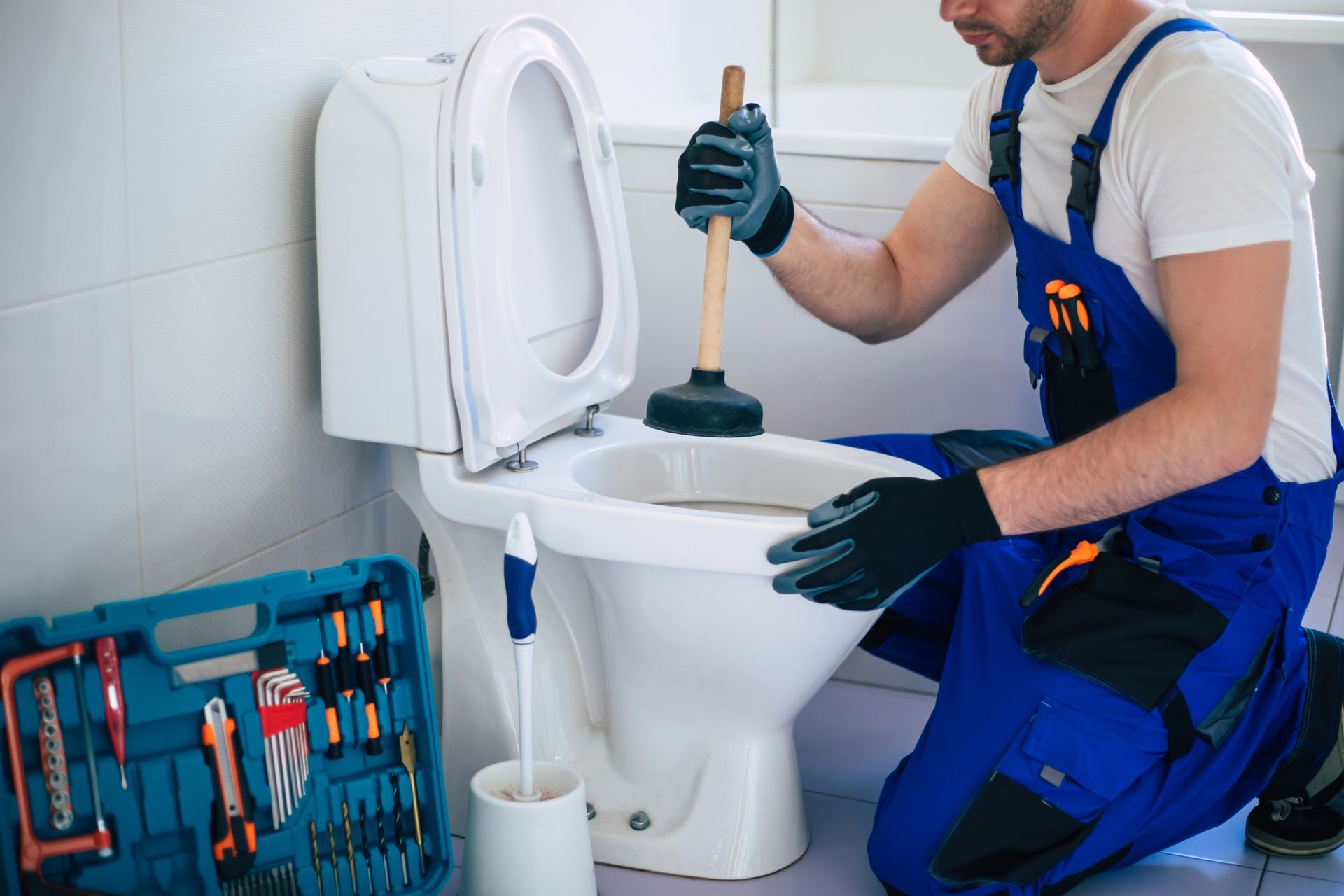 A man is kneeling down in a bathroom using a plunger to unblock a toilet.