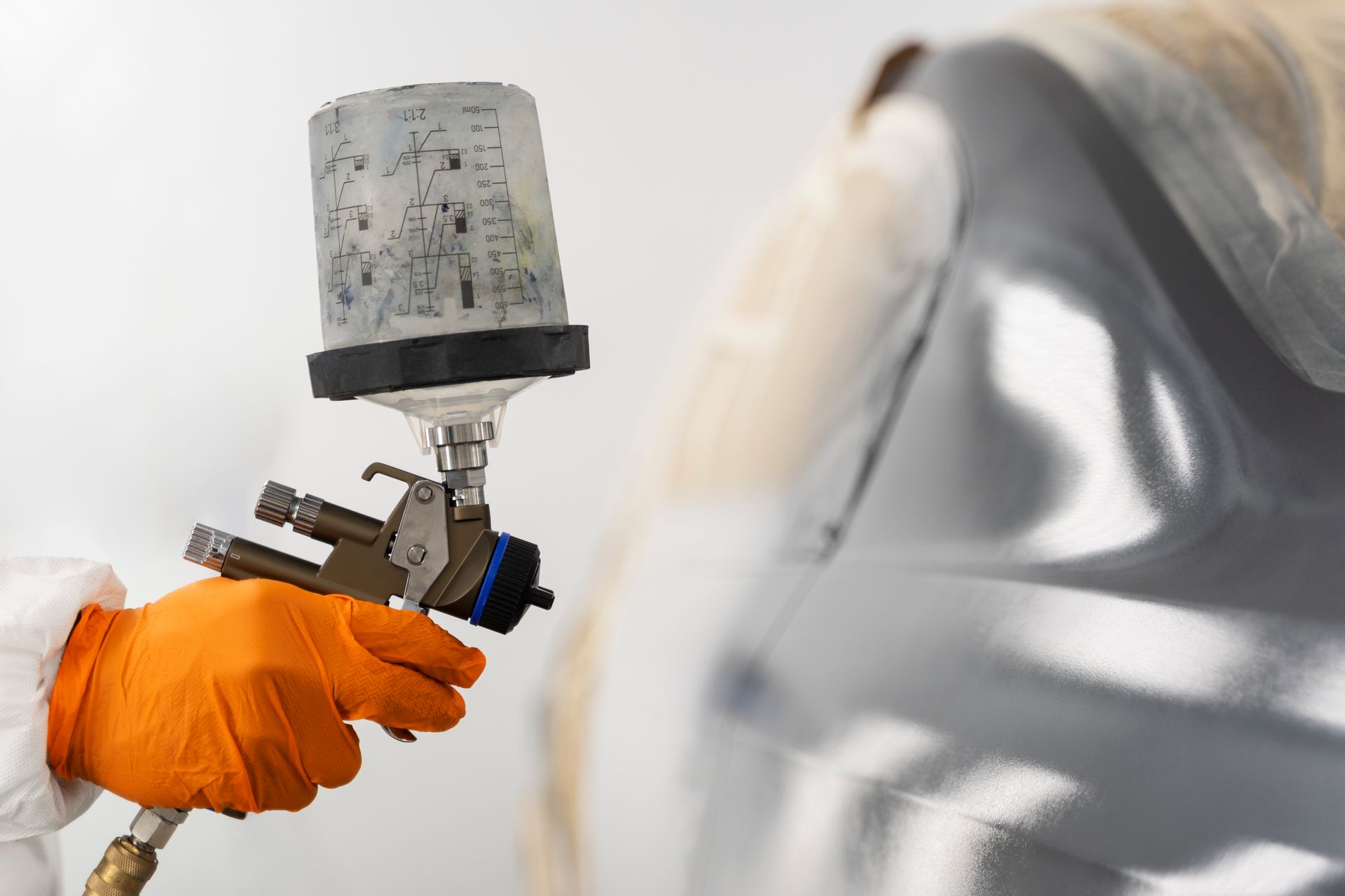 Close-up of a person in orange gloves using a spray gun to paint a car panel.