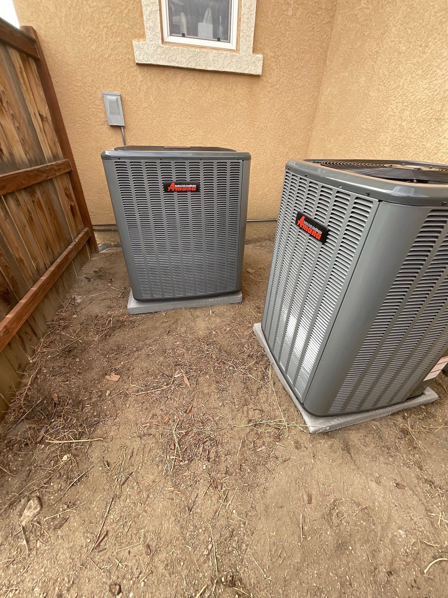 Two gray air conditioning units outside, near a fence and a building.