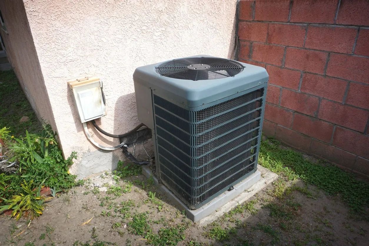 Outdoor air conditioning unit next to a wall. Gray and black, sits on a concrete pad.