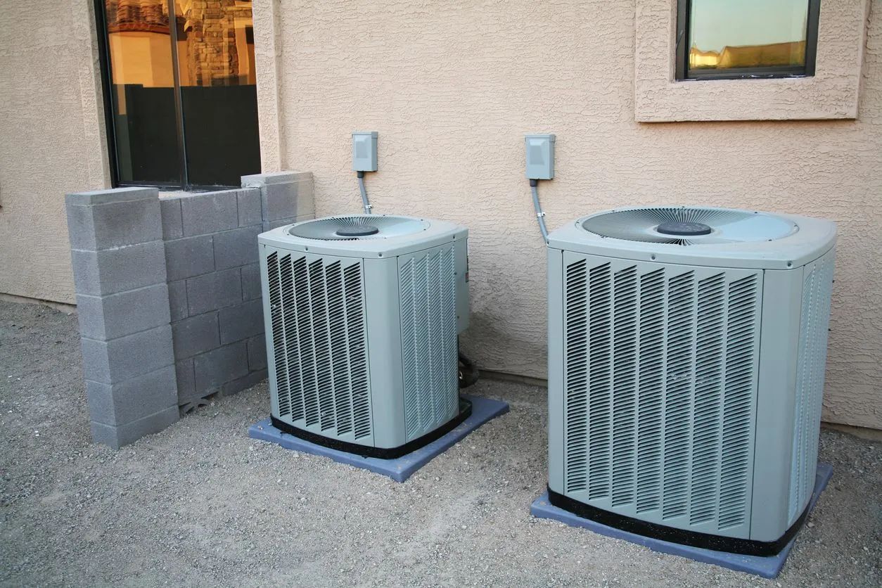 Two outdoor HVAC units beside a tan building wall on a gravel surface.