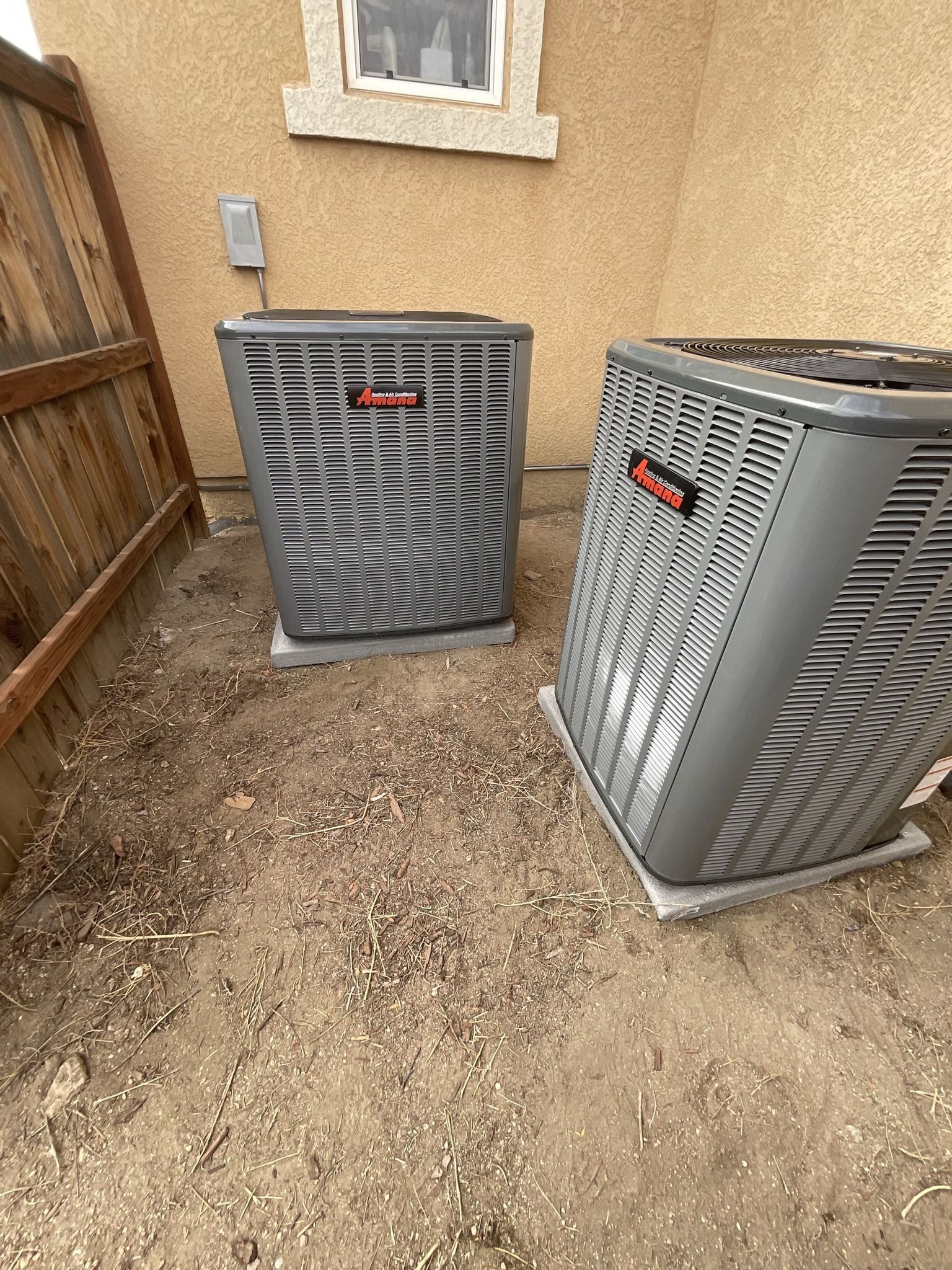 Two gray air conditioning units on concrete pads next to a fence and beige building.