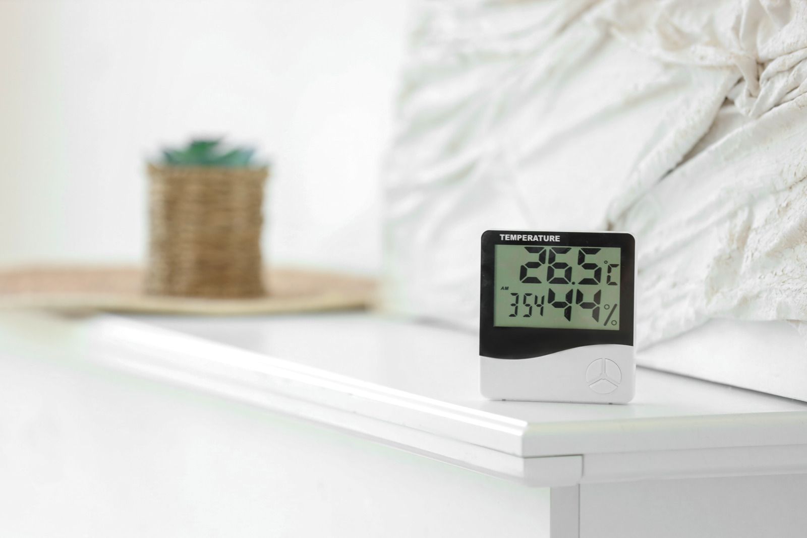 Digital thermometer on a white table in a bright bedroom, with a blurred plant and bed in the background