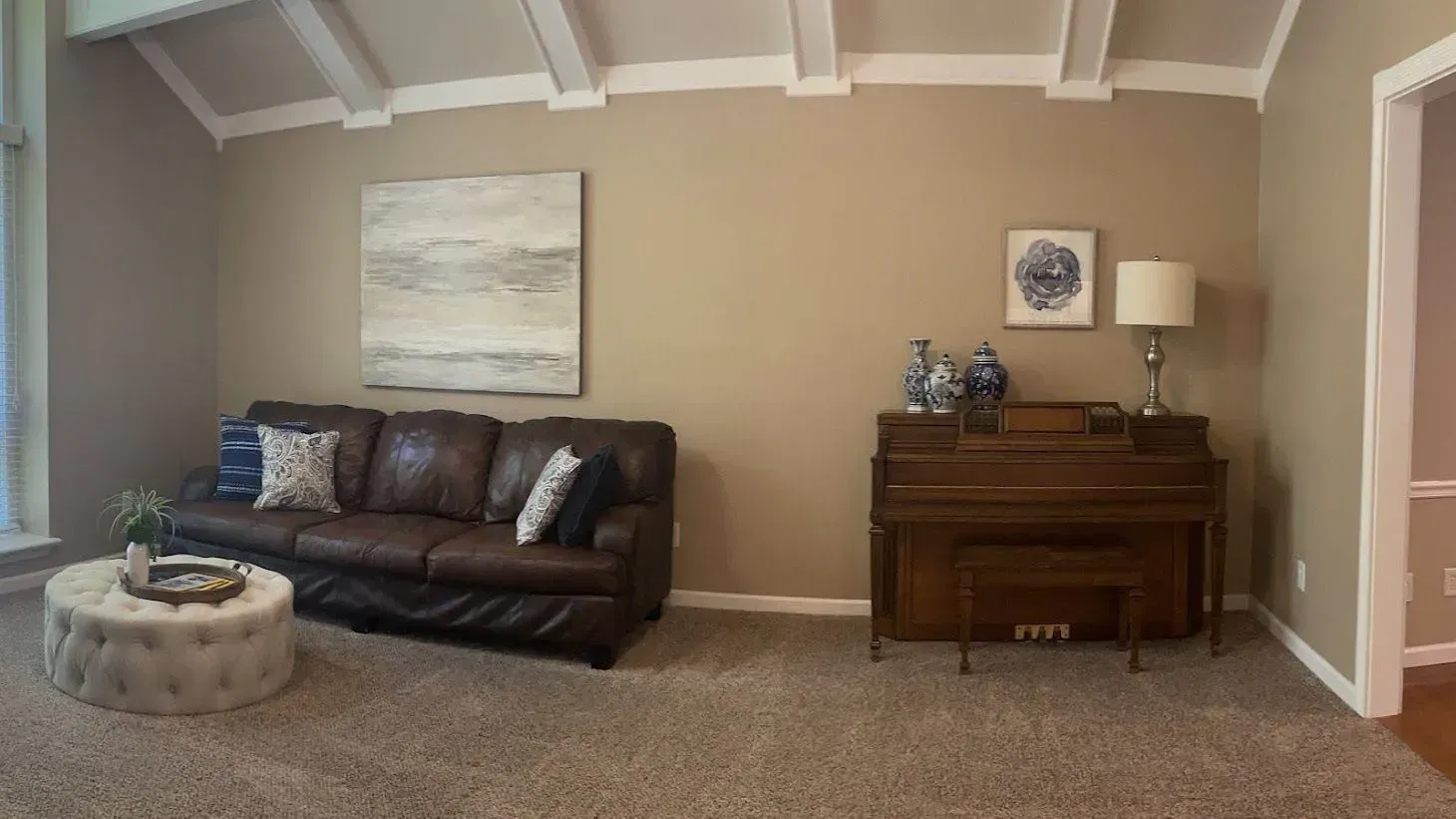 A living room with a brown leather couch, piano, and artwork on tan walls, neutral carpet, and white ceiling beams.