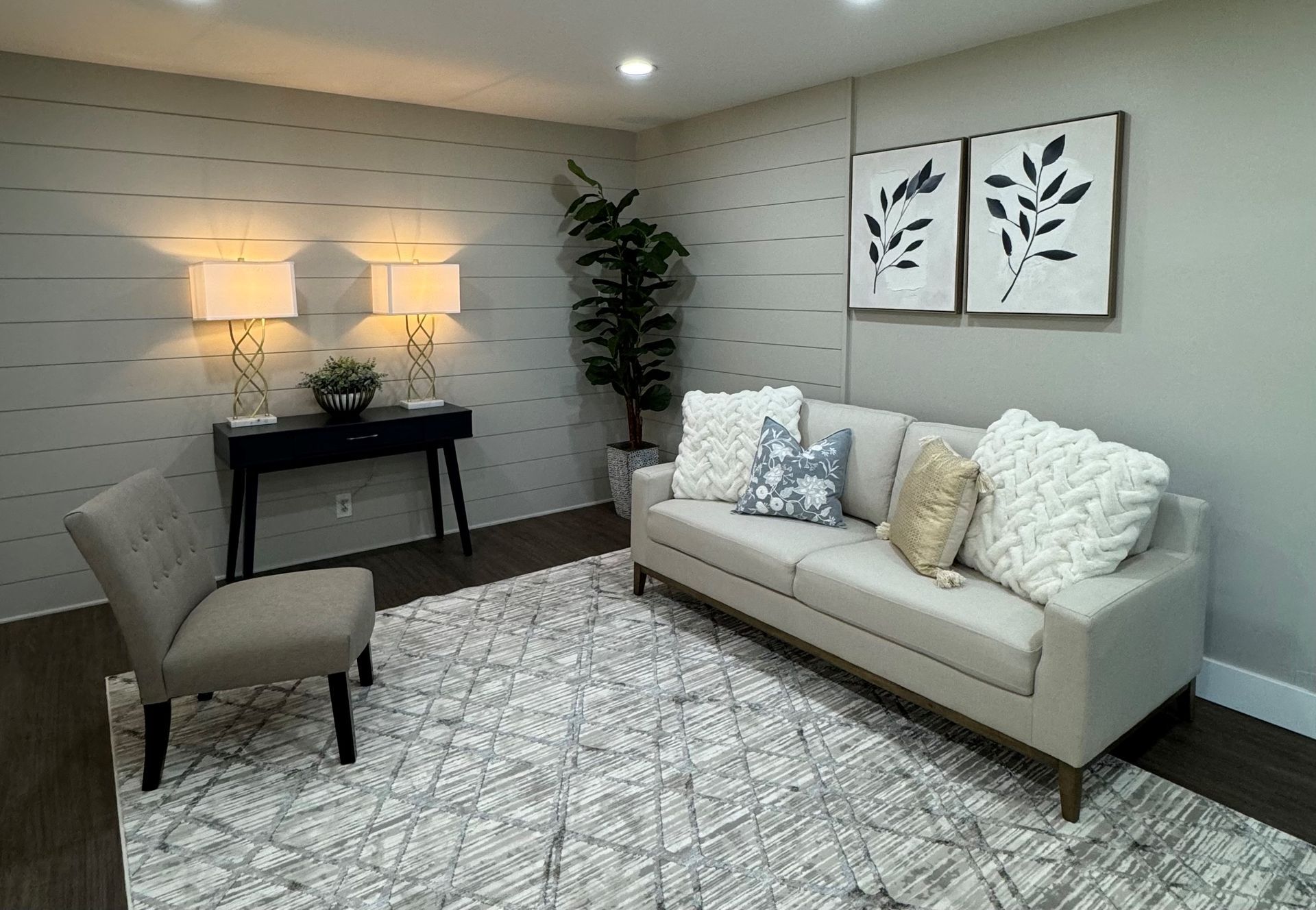 Cozy, neutral-toned living room with sofa, chair, rug, and artwork. A side table with lamps is present.