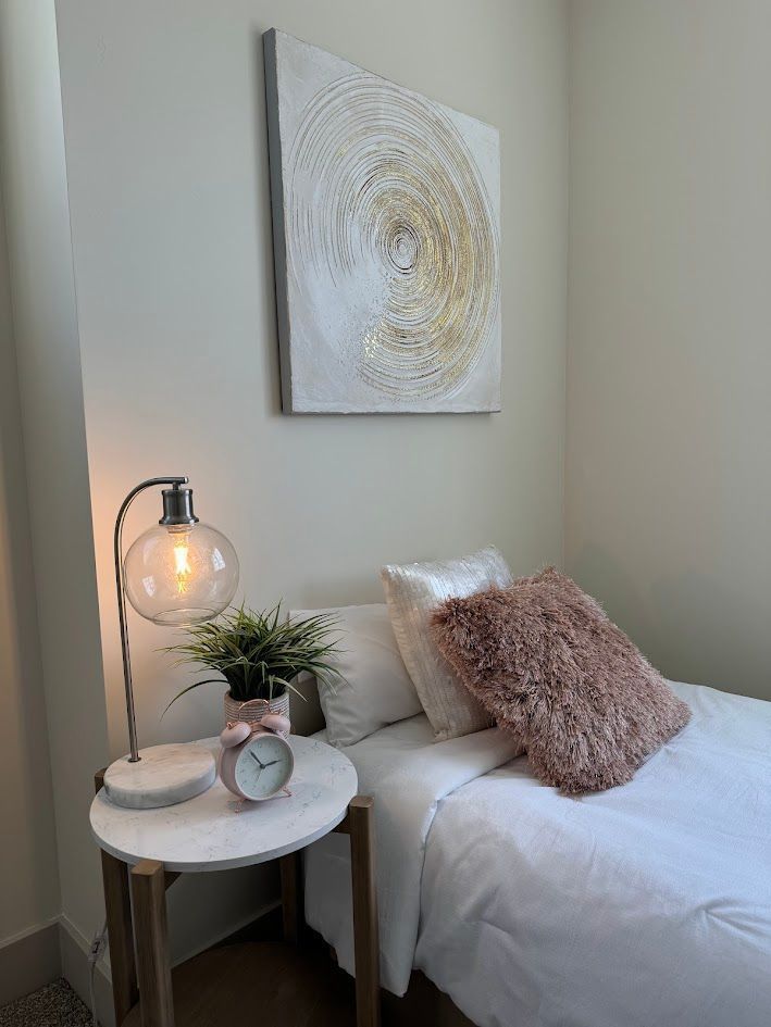 Bedroom corner with a white bed, small table with a lamp, and abstract art on the wall.