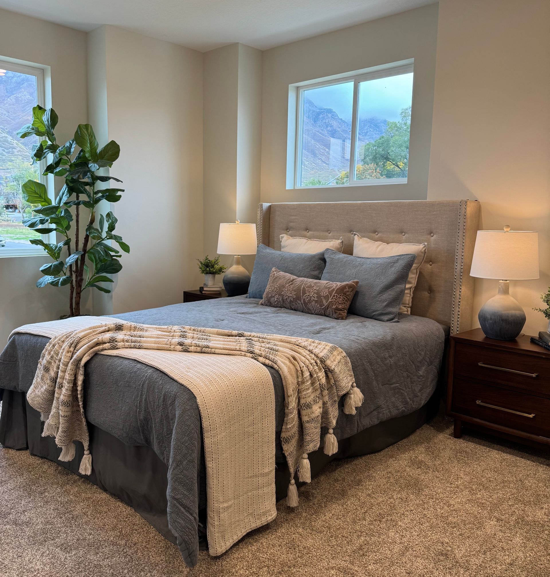 Bedroom with gray bed, nightstands, lamps, window, and tall plant.