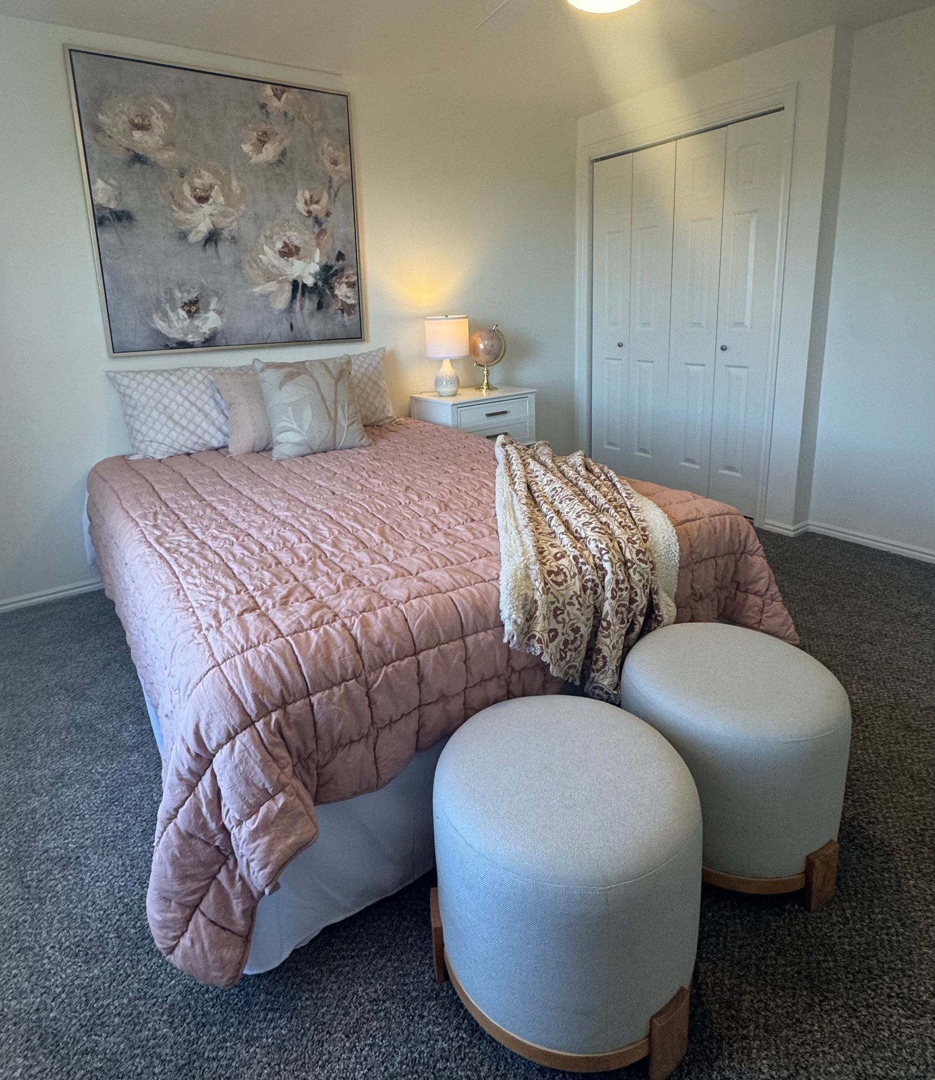 Bedroom with pink comforter, floral art, white wardrobe, two gray ottomans on a gray carpet.