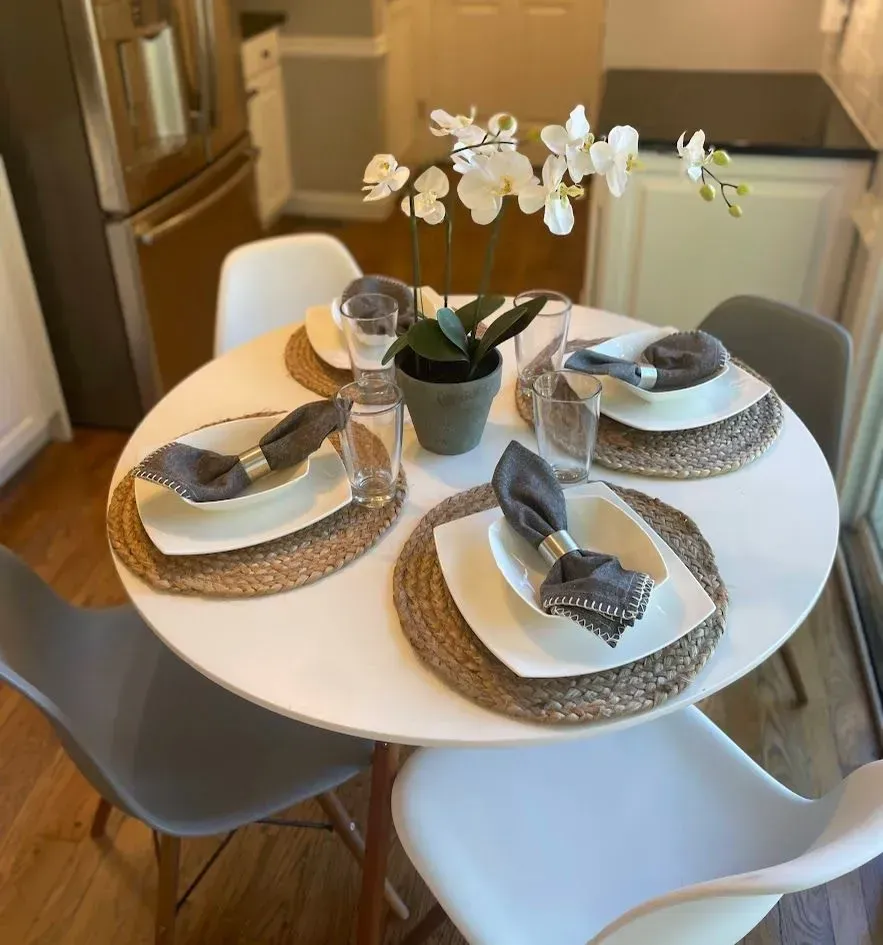 Round white table set for four, with plates, napkins, and an orchid centerpiece in a dining room.