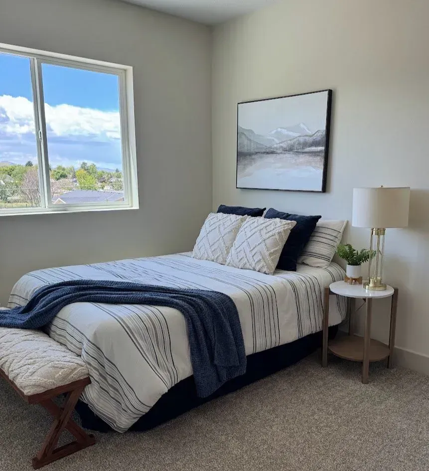 Bedroom with bed, art, and side table. Blue and white bedding, gray walls, and window with sky view.