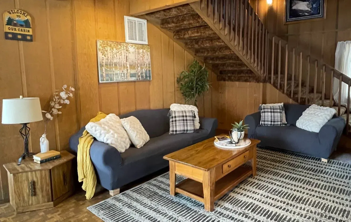 Living room with blue sofas, wooden paneling, a wooden staircase, and a striped rug.
