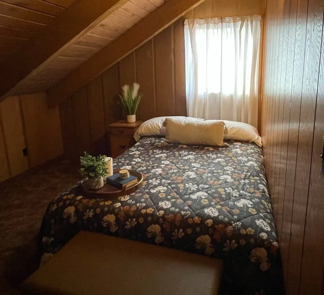 Cozy bedroom with slanted ceiling, bed with floral comforter, nightstand with plant, and window with sheer curtain.