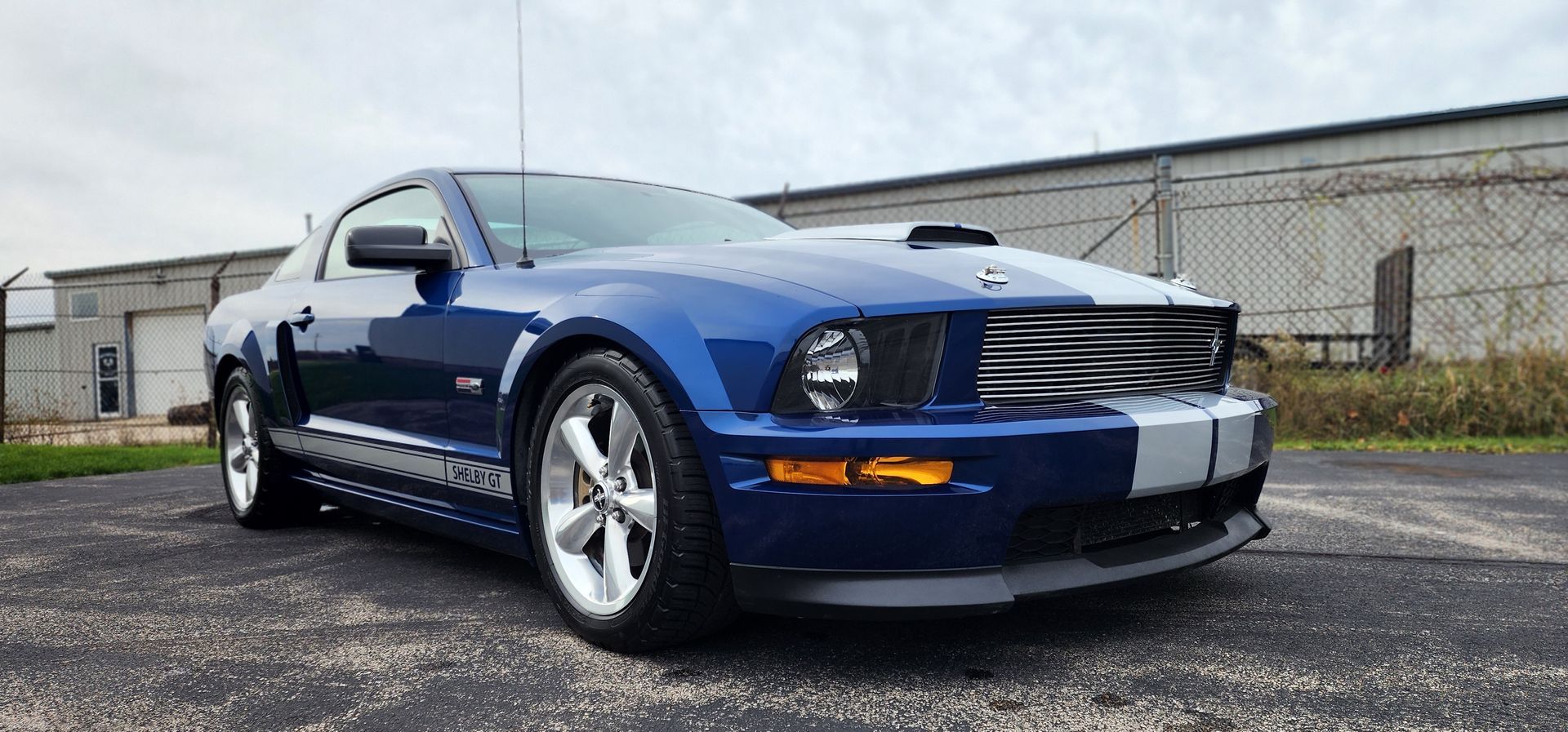 Blue Ford Mustang with white stripes in a garage next to a blue car. Tools and boxes are present.