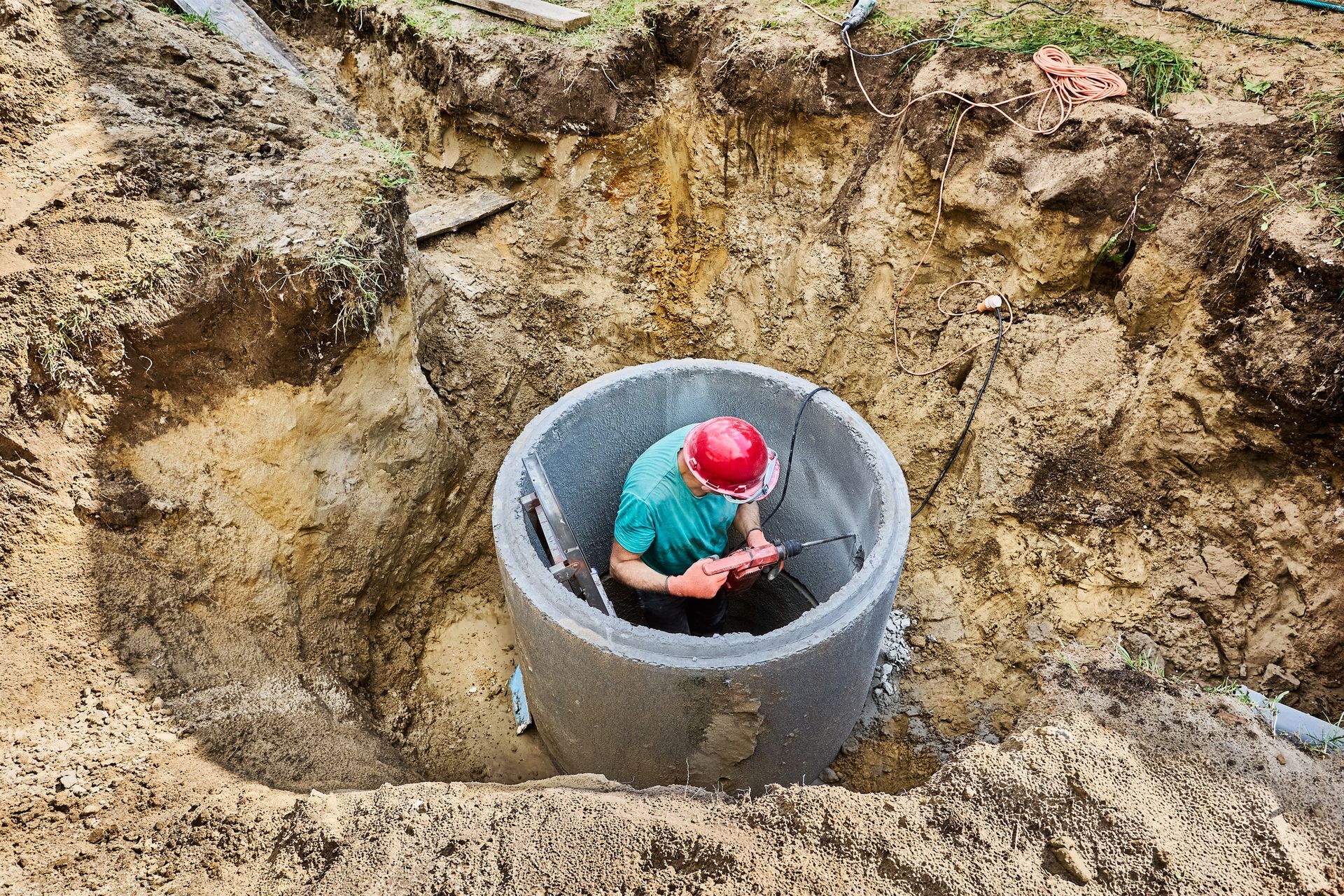 A person wearing a red helmet works inside a concrete well, dug in brown earth.