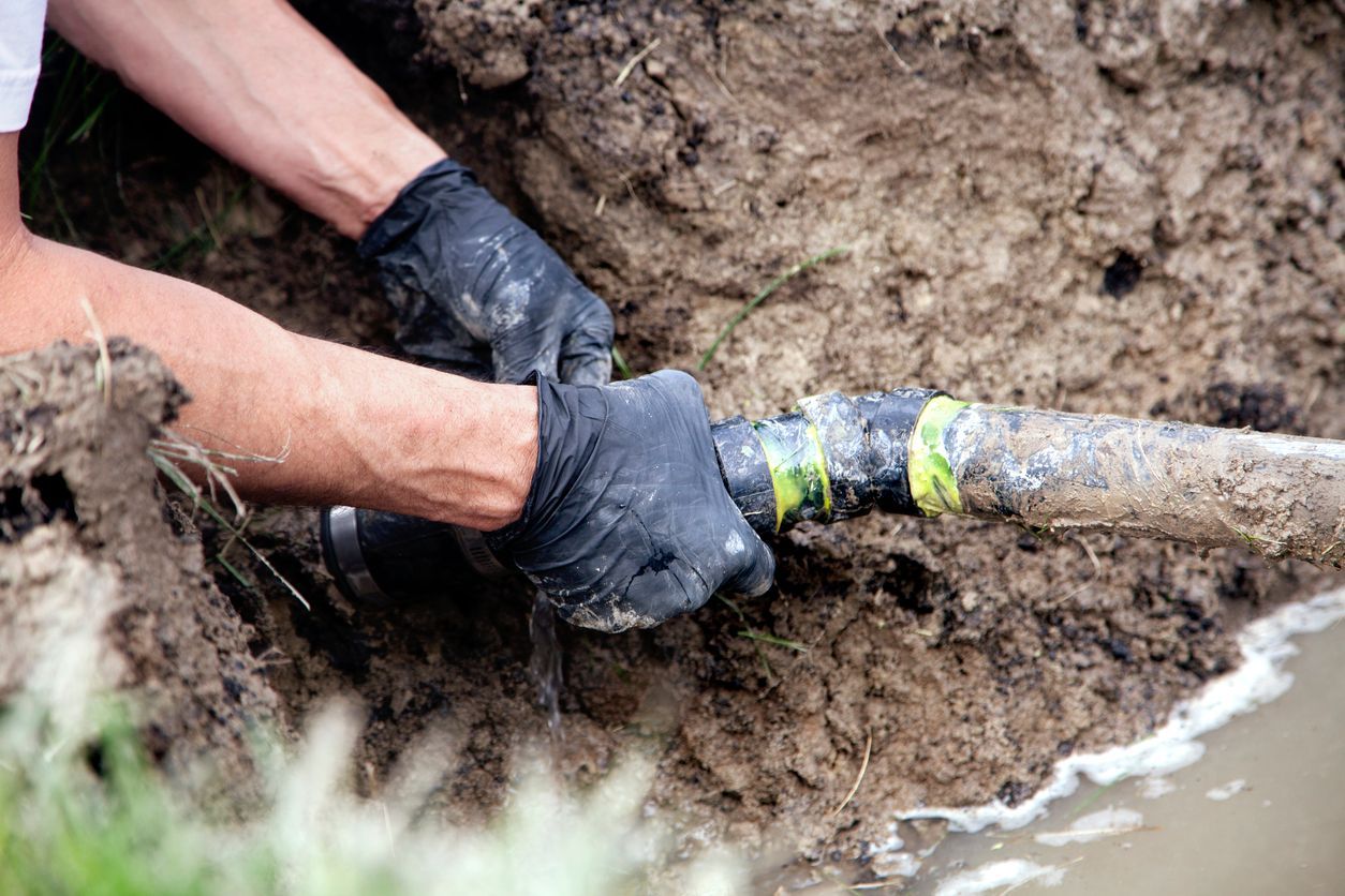 Hands in black gloves repairing a cracked pipe in a dirt trench.