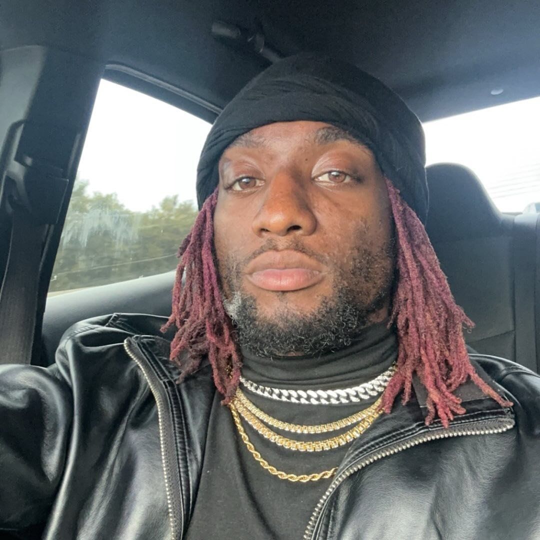 Man in black beanie and jacket with red dreads, looking at the camera.