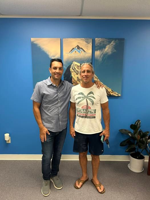Two men standing in front of a blue wall with art, one in a shirt and jeans, the other in a t-shirt and shorts.