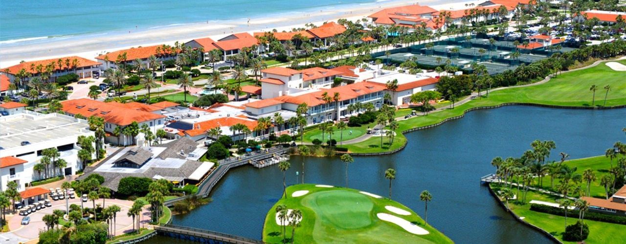Aerial view of a coastal resort with buildings, tennis courts, golf course, and a sandy beach.