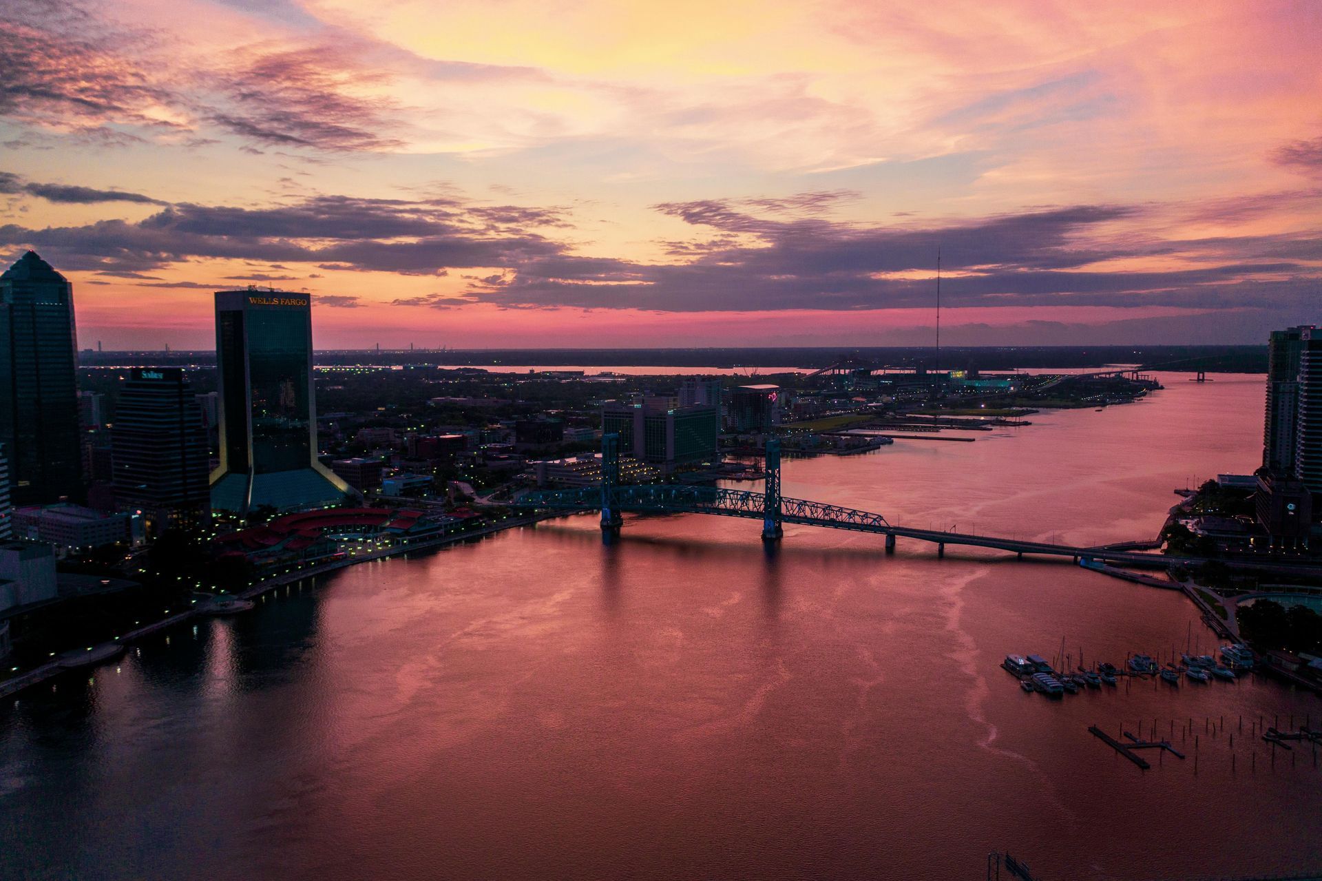 Sunset over Jacksonville, Florida, with a bridge spanning a wide river reflecting the colorful sky; city skyline in view.