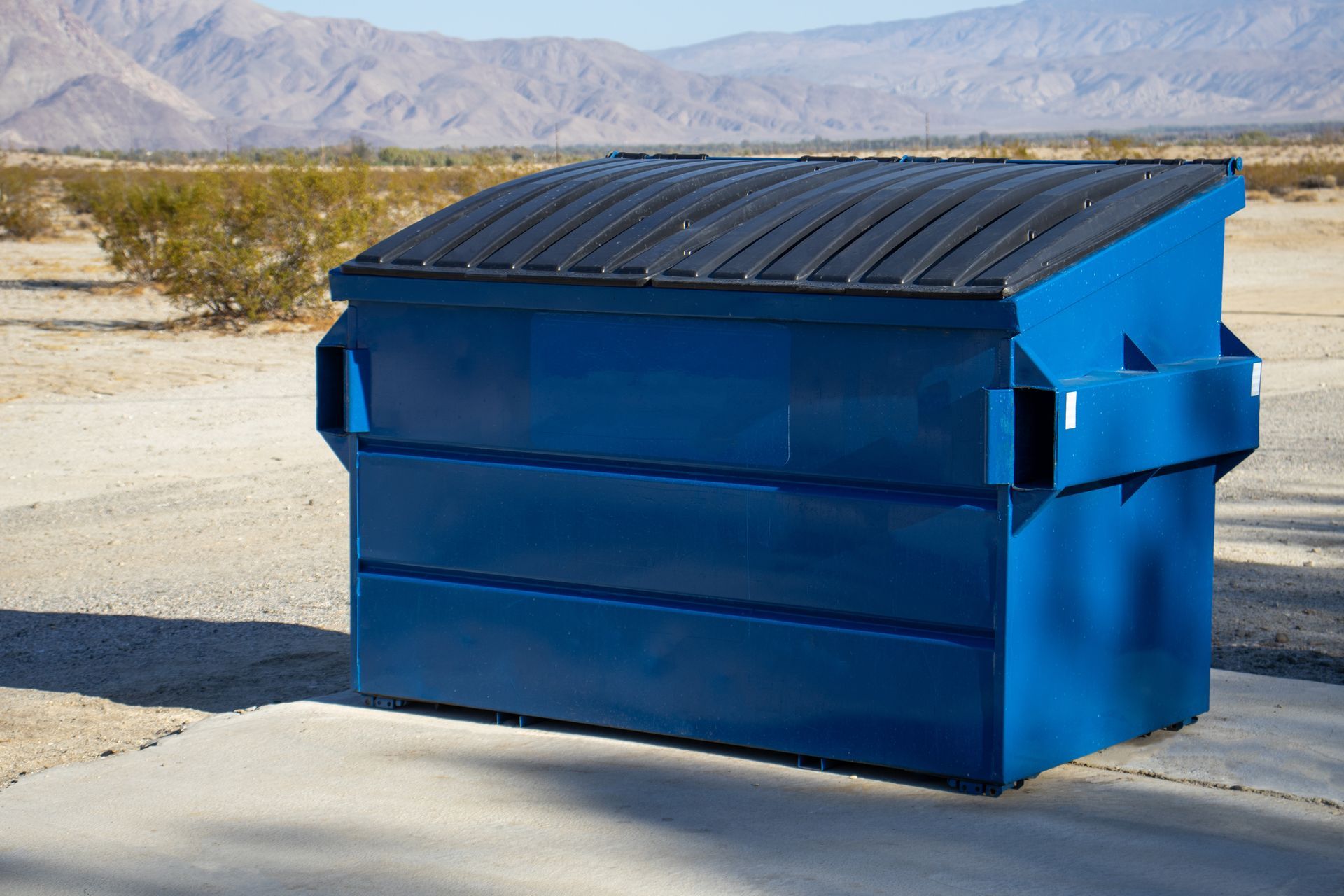 Large blue metal dumpster with a closed lid placed on a concrete pad in a desert area. Large blue metal dumpster with a closed lid placed on a concrete pad in a desert area.