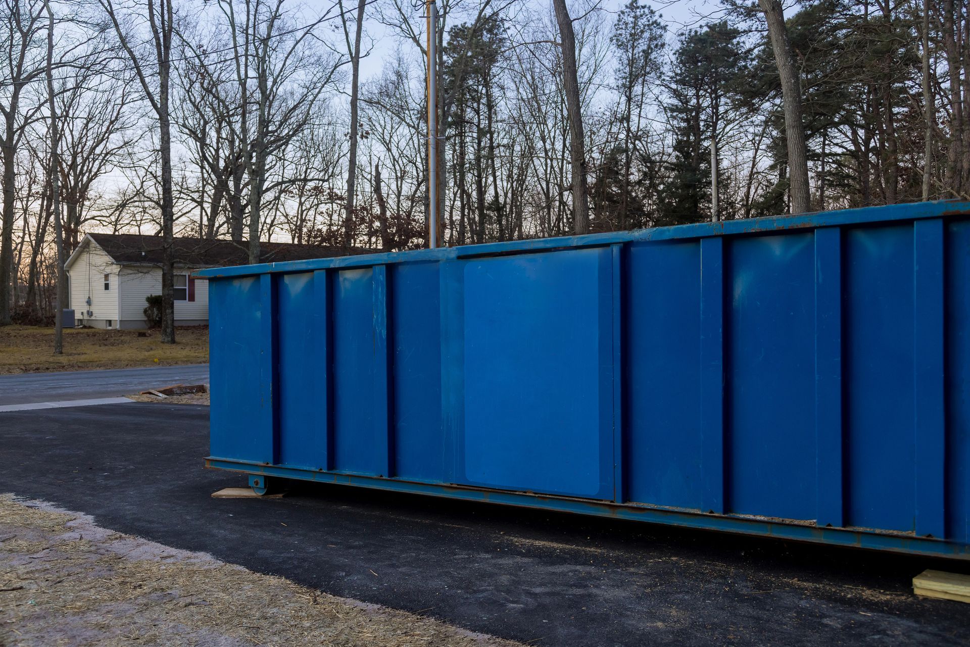 A blue dumpster is sitting in a parking lot with trees in the background.