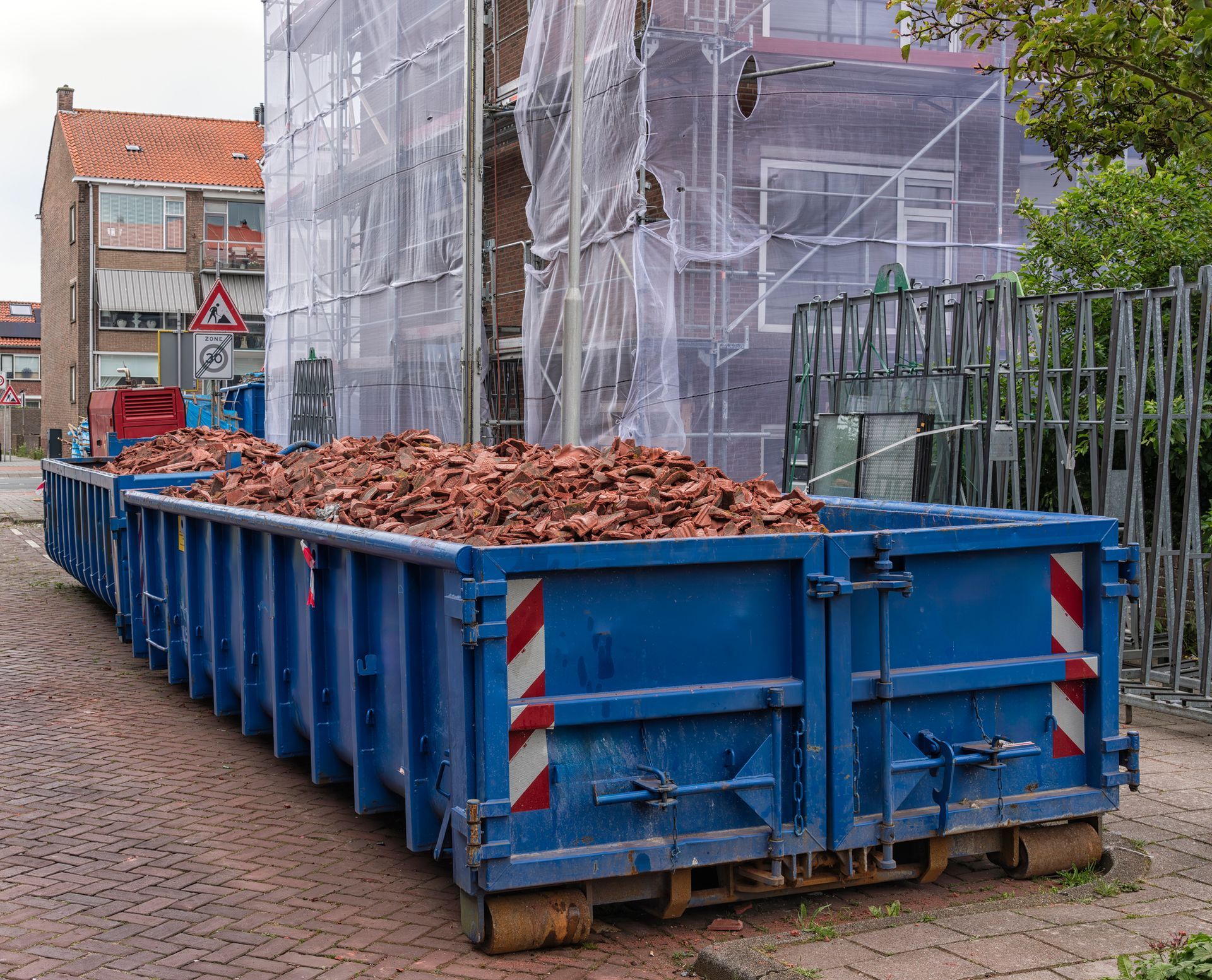 A blue dumpster filled with bricks is parked in front of a building under construction.