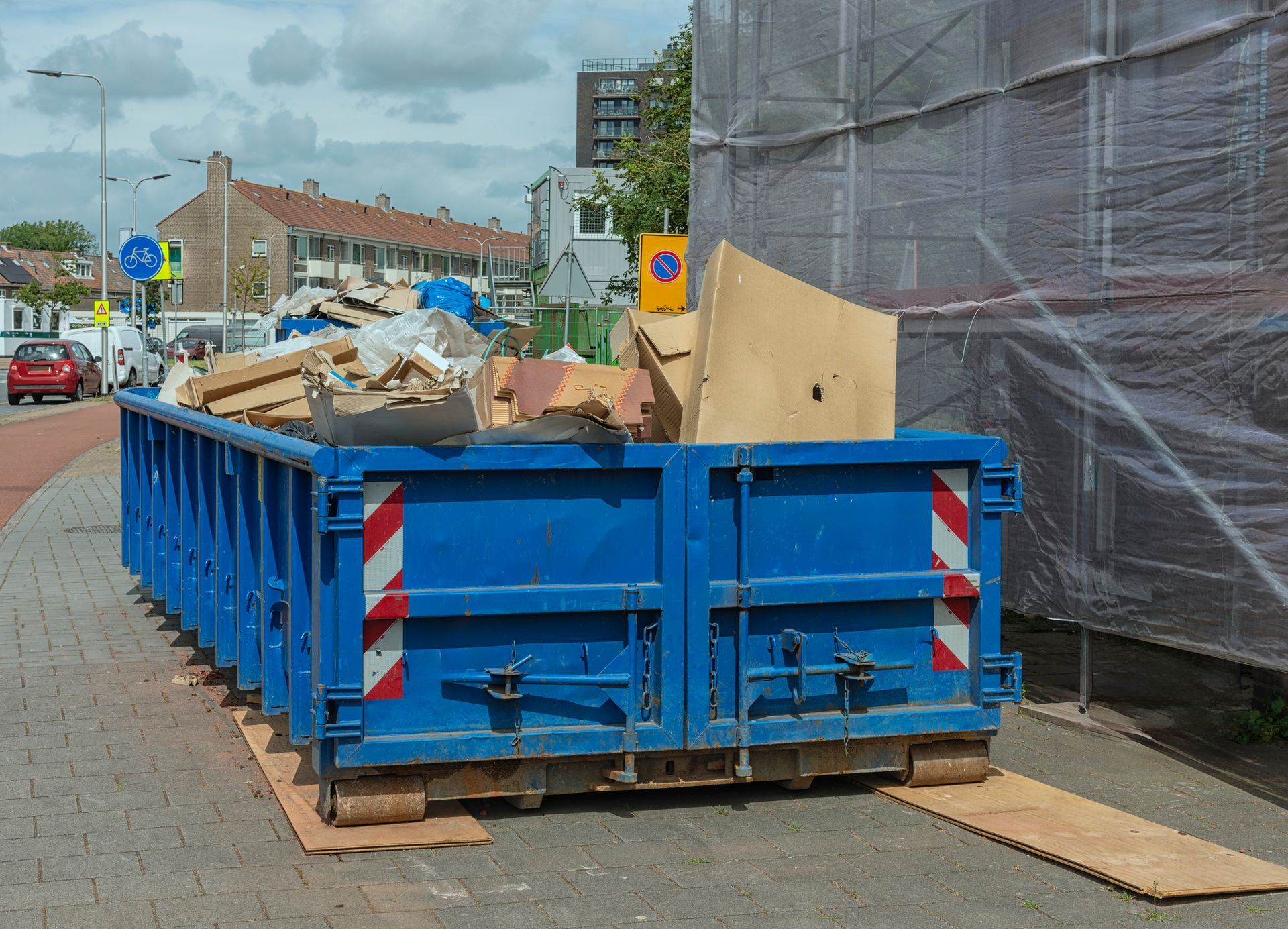 Blue steel dumpster filled with cardboard waste placed on a sidewalk in a residential neighborhood.