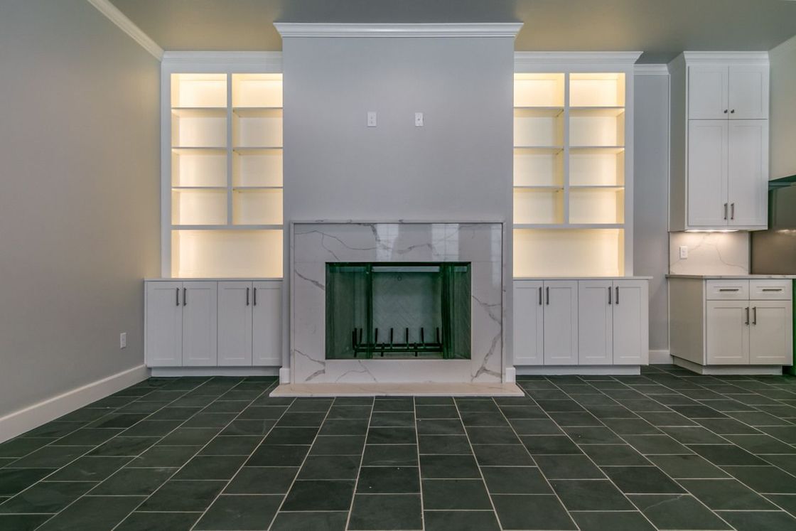 Living room with fireplace, built-in shelves, white cabinets, marble, and gray walls. Dark stone floor.