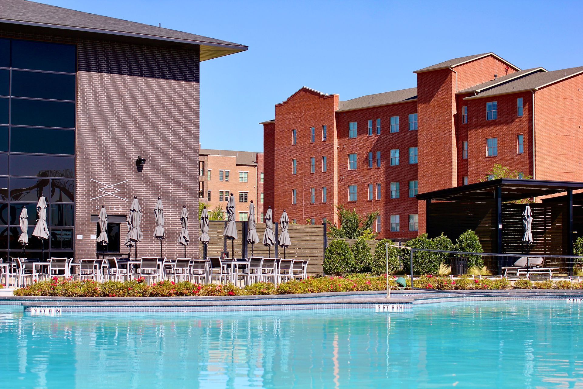 Swimming pool in front of brick buildings on a sunny day. Tables with umbrellas, blue sky.