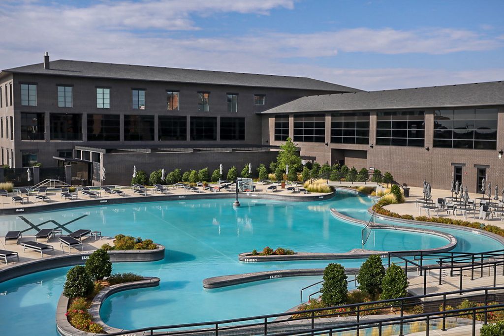 Hotel with a large, winding blue pool, surrounded by lounge chairs and greenery. Building is gray with large windows.