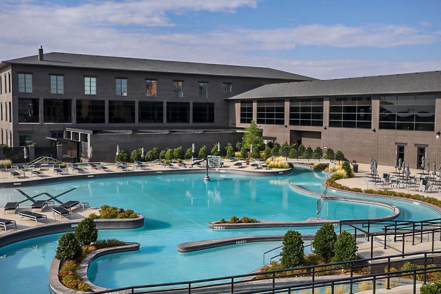 Hotel with a large, winding blue pool, surrounded by lounge chairs and greenery. Building is gray with large windows.