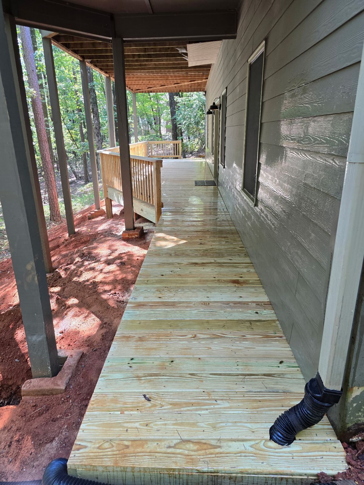 Wooden porch with railing and columns, next to a house with a rain gutter, in a wooded setting.