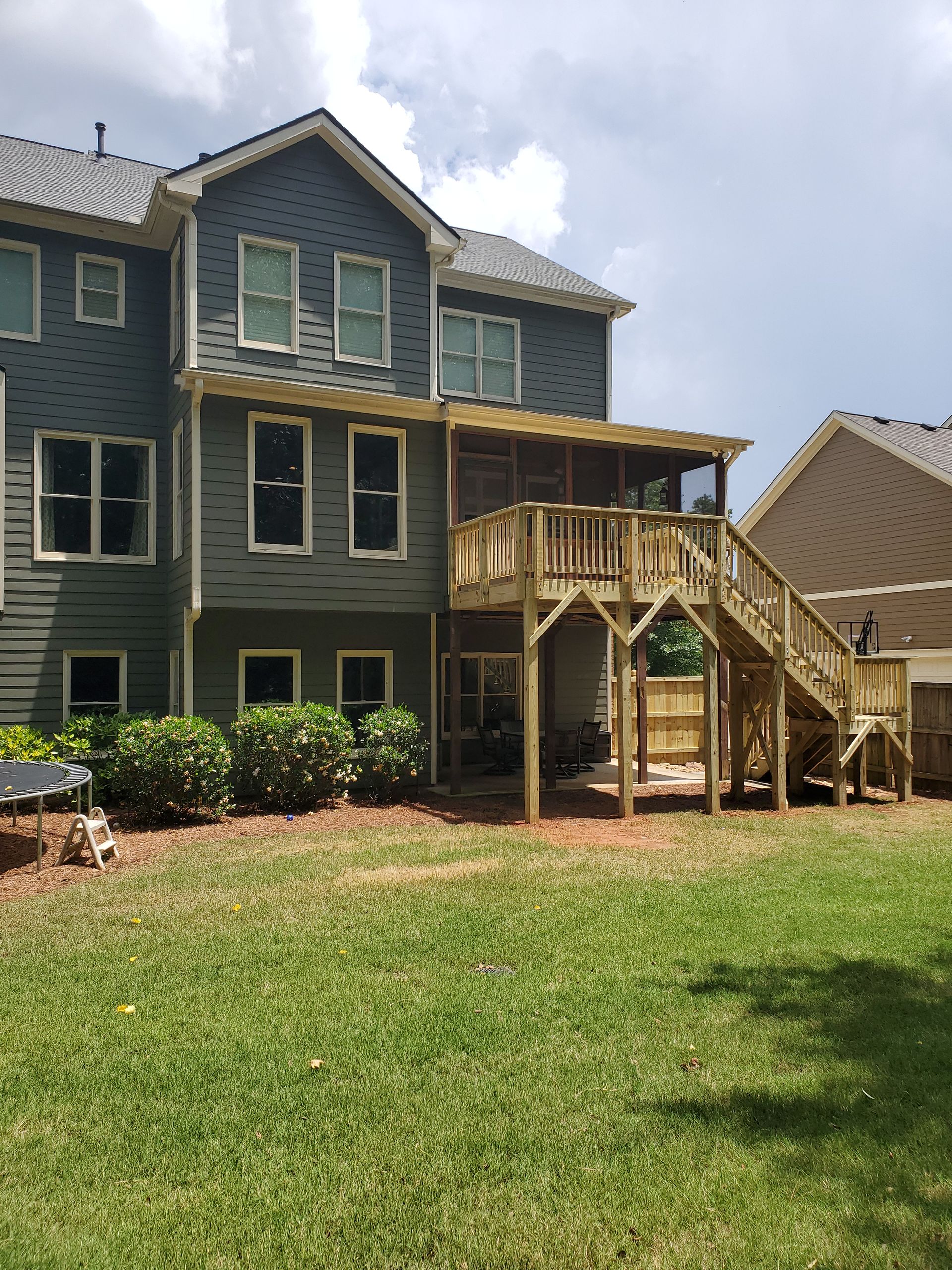 Rear view of a two-story home with a wooden deck and staircase leading to a screened porch. Green grass and bushes in front.