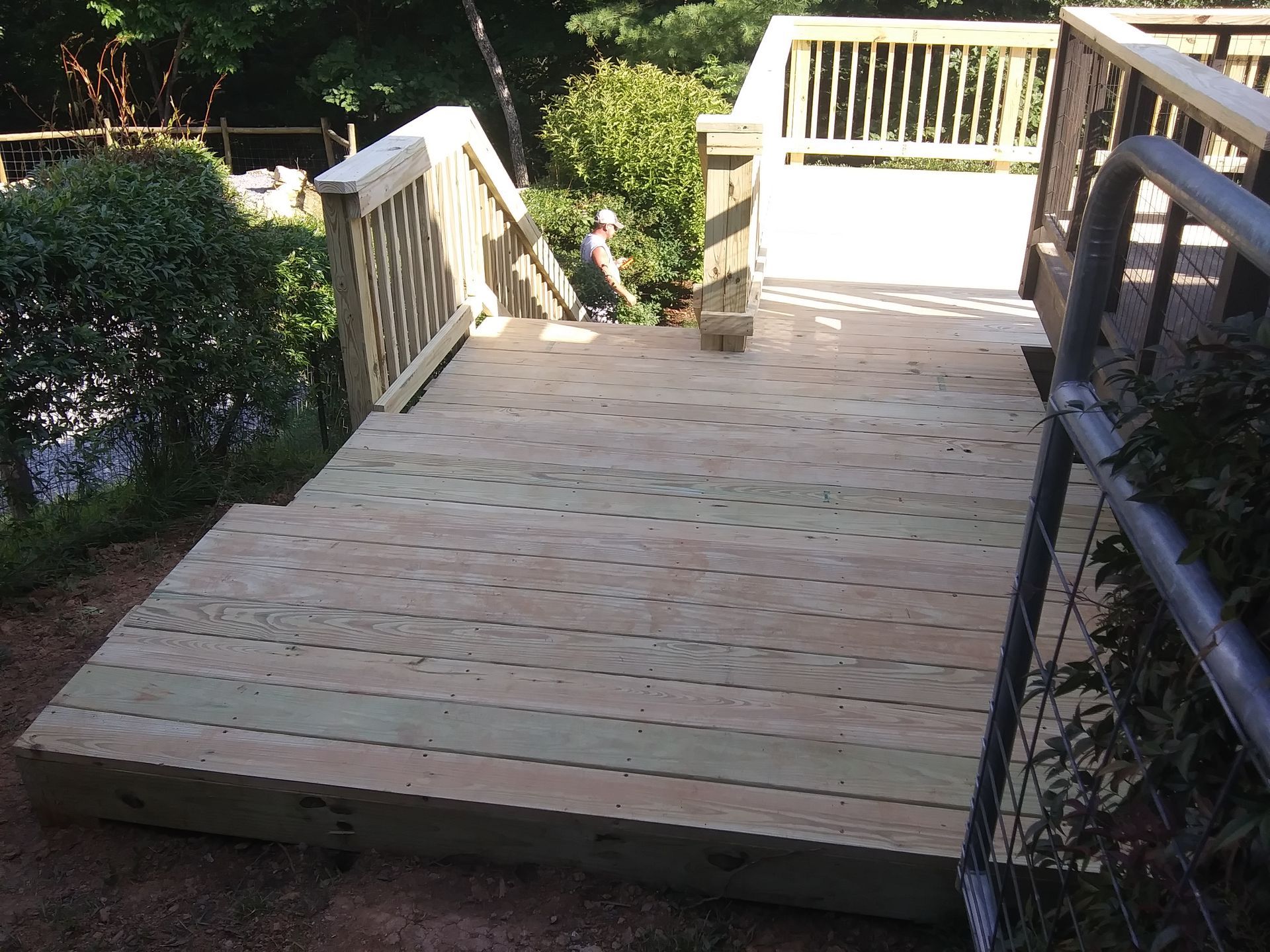 Wooden deck with stairs, railing, and a gate. Brown wood, outdoor setting with greenery.