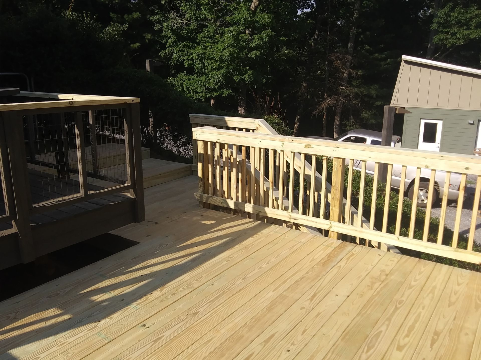 Wooden deck with railing. Light brown wood, sunlight, and a house in the background.