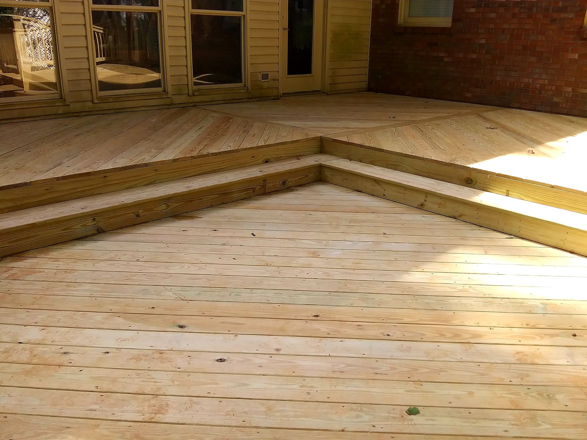 Wooden deck with two steps leading down. A house and brick wall are in the background.
