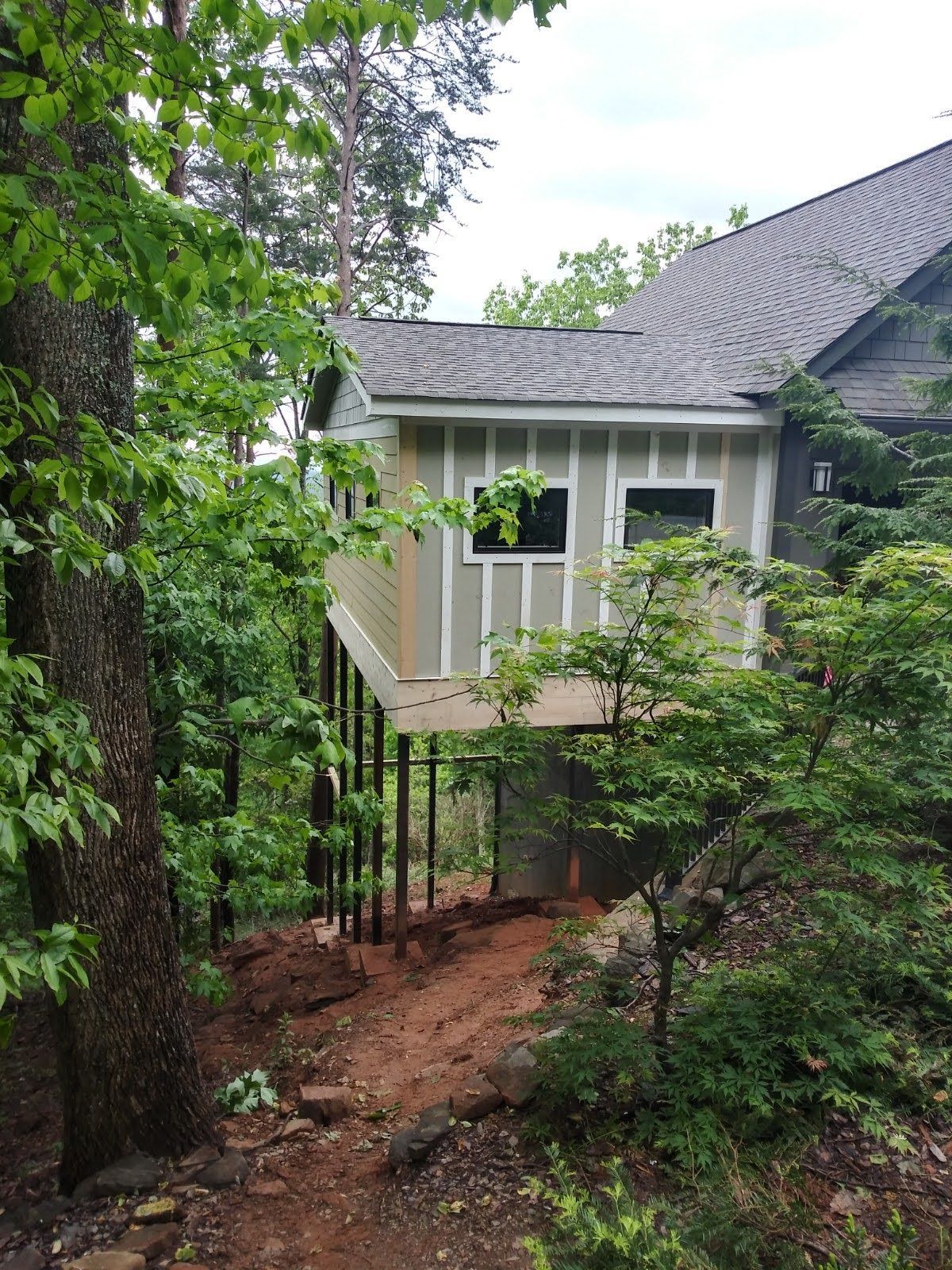Elevated structure with light-colored siding, dark roof, supported by poles, set in a wooded area with trees.