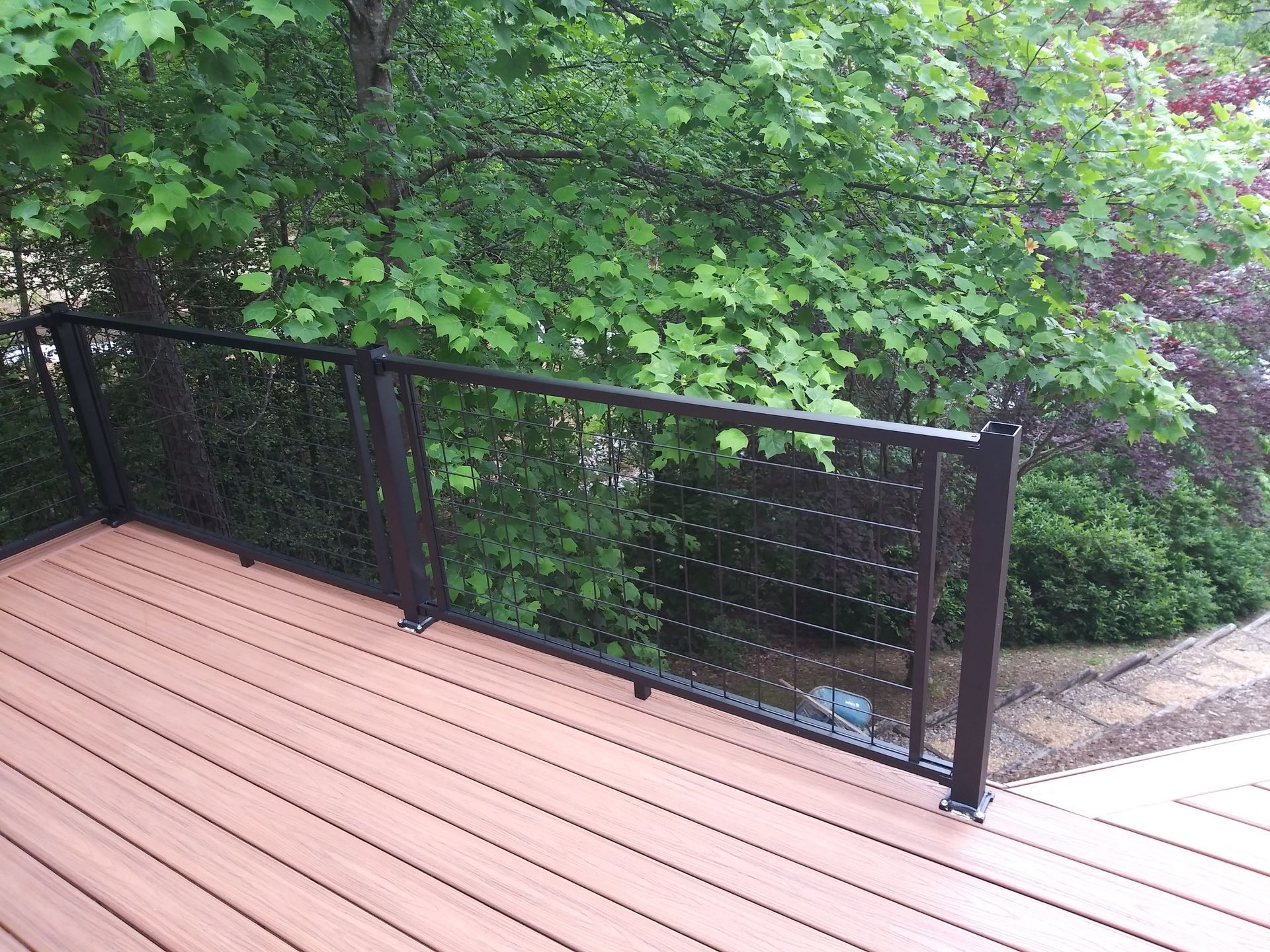 Deck with black metal railing and cable infill, overlooking green foliage. Brown deck boards.