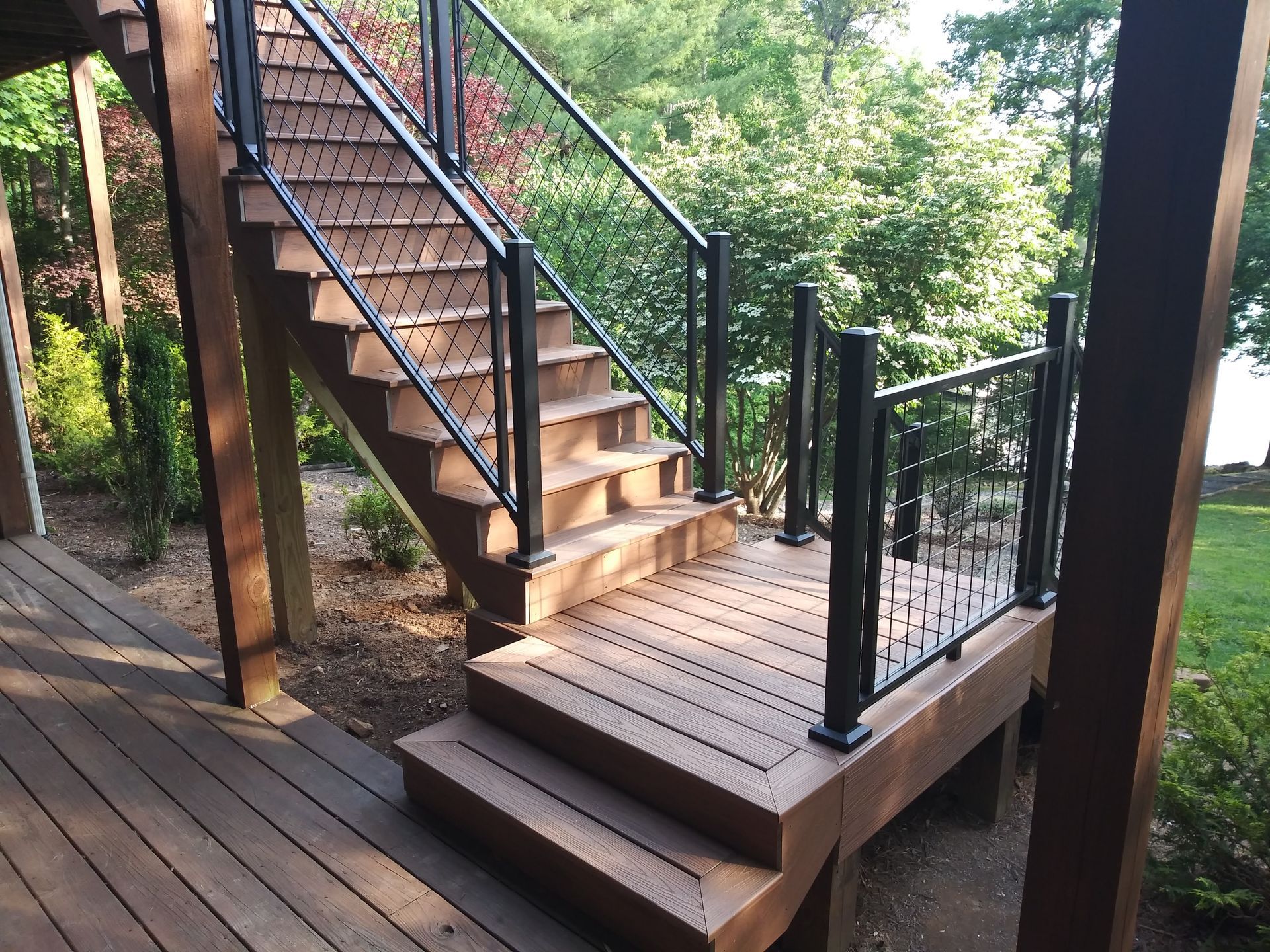 Wooden deck stairs with black railings leading to a deck, surrounded by greenery.