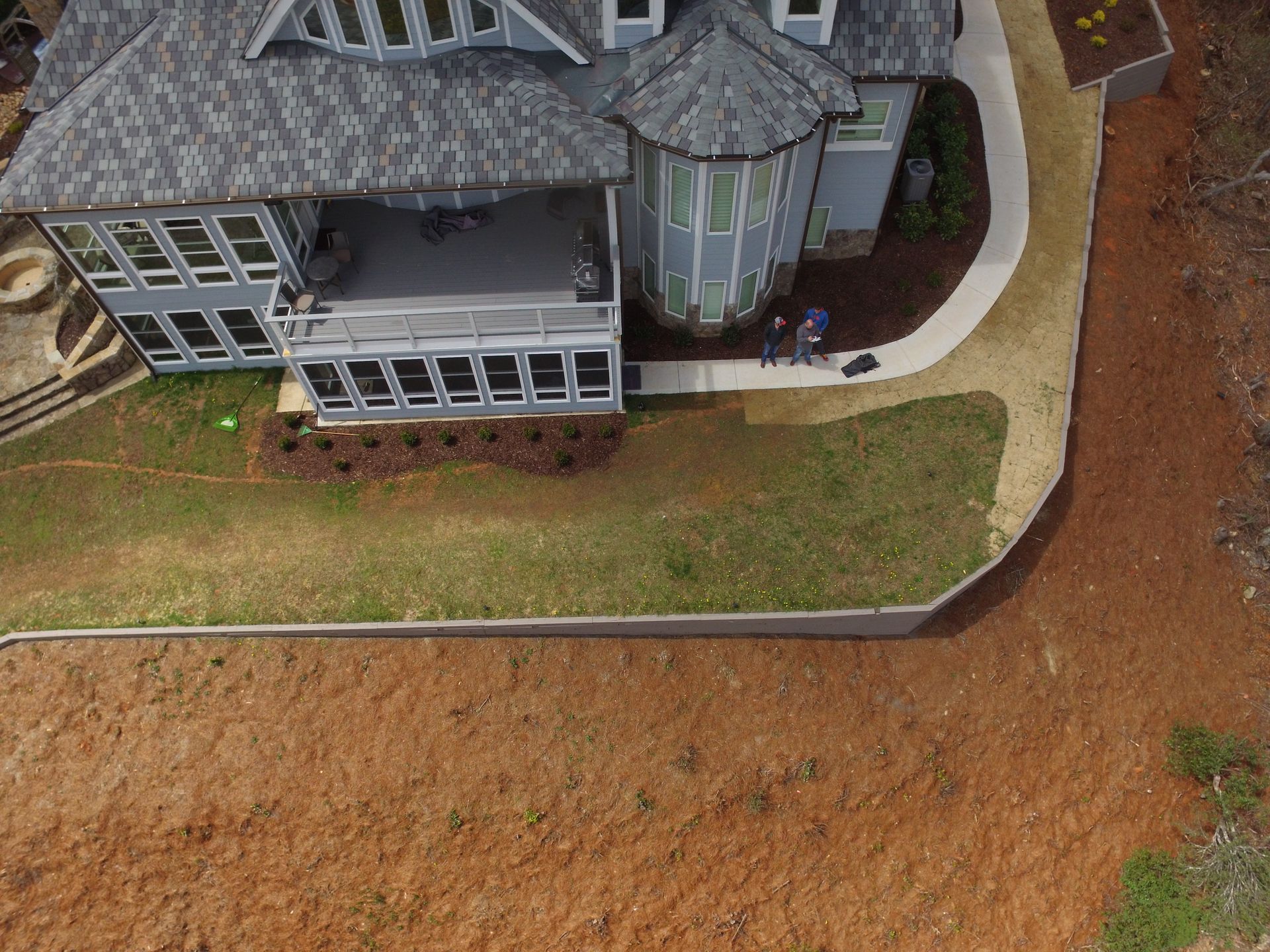 Aerial view of a gray house with a curved front. A walkway leads to the entrance. Brown mulch and red earth are visible.