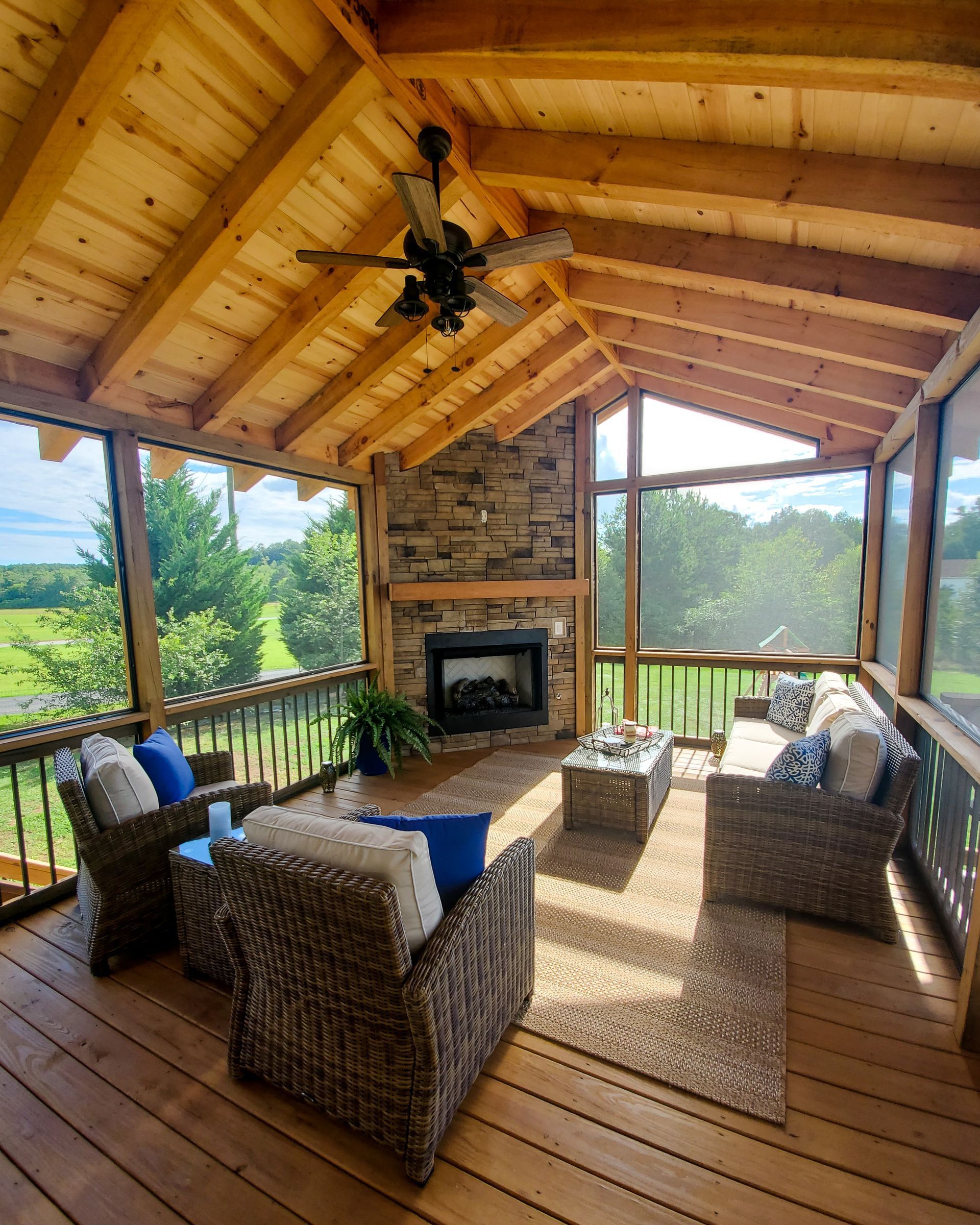 Screened-in porch with fireplace, seating, and wooden ceiling. Views of green fields, sunny day.
