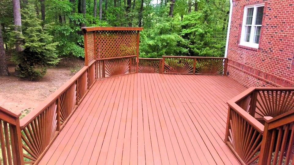 Wooden deck with brown railings, adjacent to a brick house and surrounded by trees.