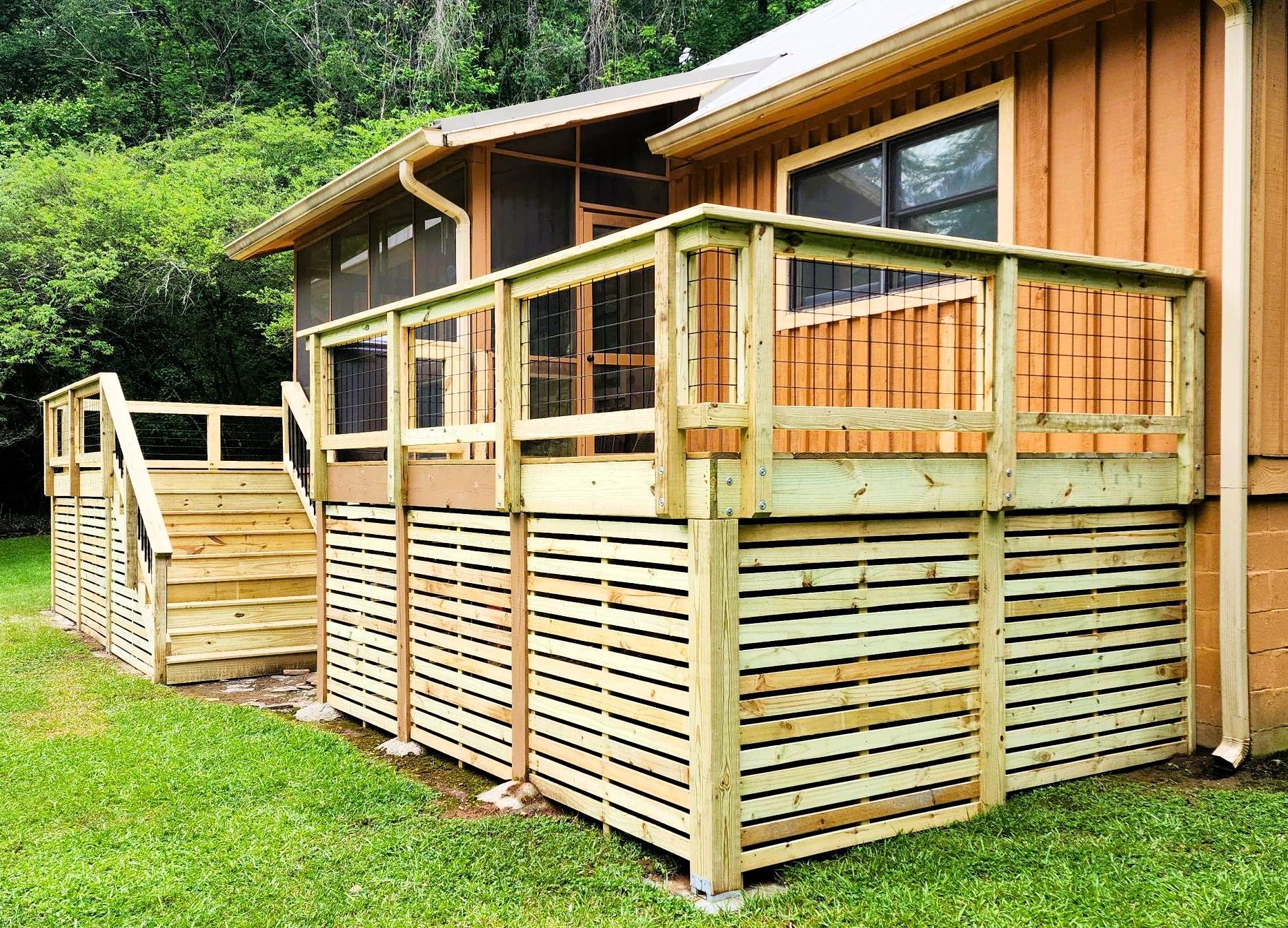 Wooden deck with stairs and lattice skirting, attached to a brown-sided house, surrounded by grass.