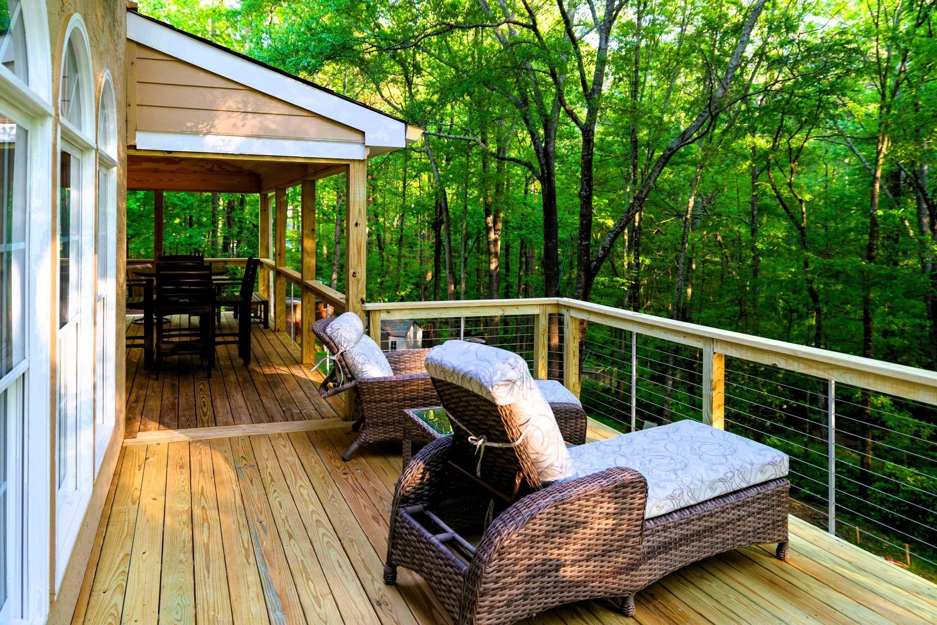 Wooden deck with lounge chairs overlooking a lush green forest. Covered porch with table and chairs.