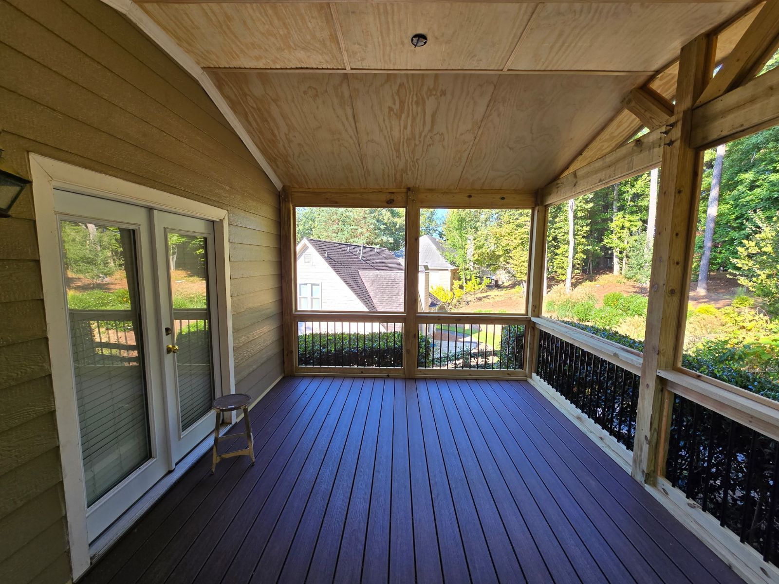 Screened porch with dark wood deck, glass doors, and view of trees and a house.