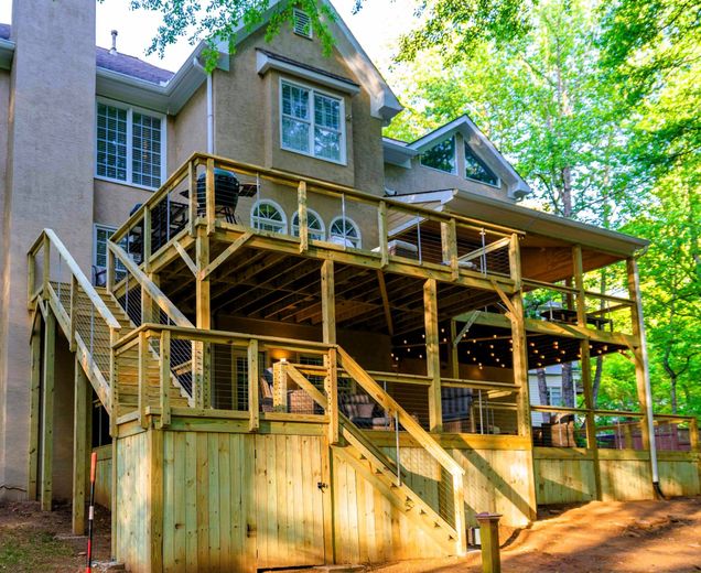 Multi-level wooden deck attached to a light-colored house, with stairs, railings, and string lights, surrounded by trees.