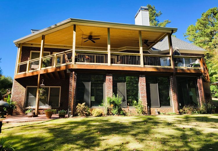 Two-story brick house with a wooden deck and covered porch. Green lawn and trees surround the house on a sunny day.