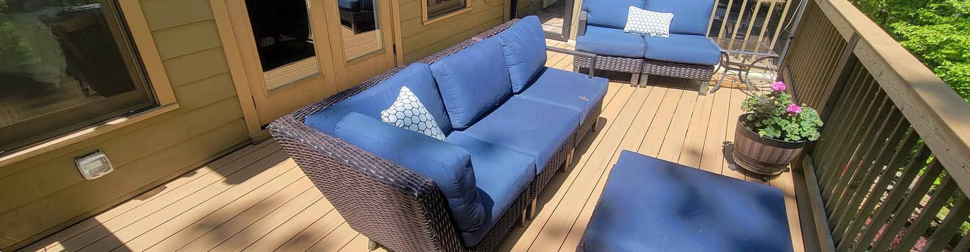 Blue outdoor furniture on a wooden deck. A three-seat sofa, a loveseat, and an ottoman are visible. Potted plants on the side.