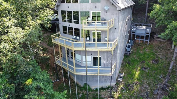 Multi-story house on a hillside with decks; gray siding, surrounded by trees.
